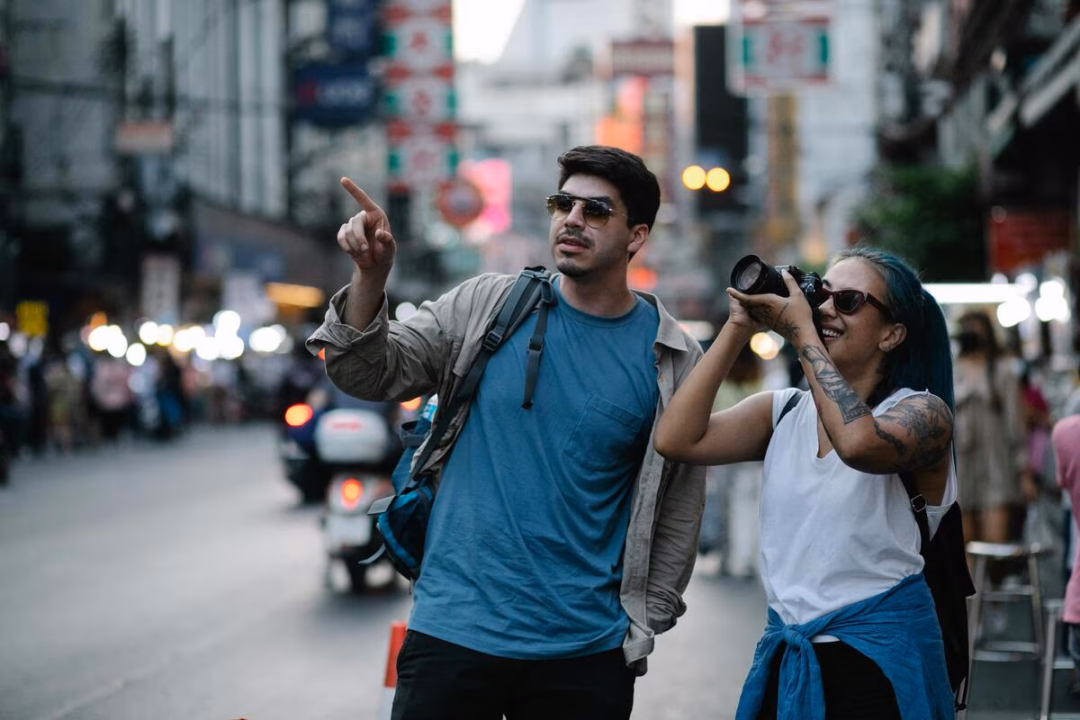 Man pointing while woman with tattoos takes a photo on a busy city street at dusk.