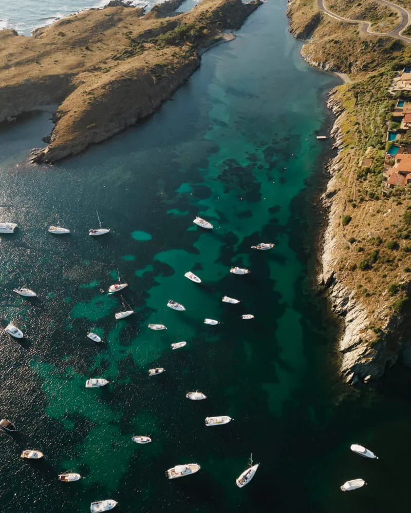 Aerial view of boats anchored in clear turquoise water along a rocky coastline with dry hills and houses.