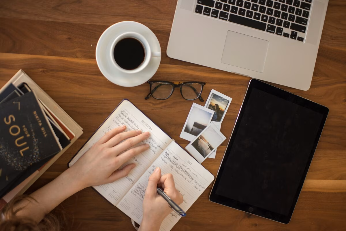 Person writing in a planner on a wooden table with a laptop, tablet, coffee cup, glasses, and photos nearby.