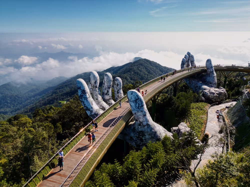 Curved pedestrian bridge supported by two giant stone hands over lush green hills under a partly cloudy sky.