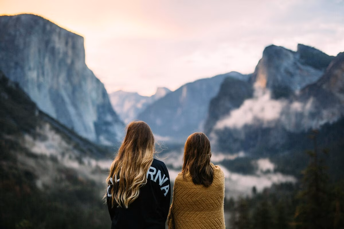 Two women with long hair looking at a misty mountain valley during sunrise or sunset.