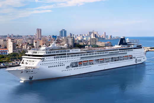Large white MSC cruise ship sailing near a city coastline under a partly cloudy sky.