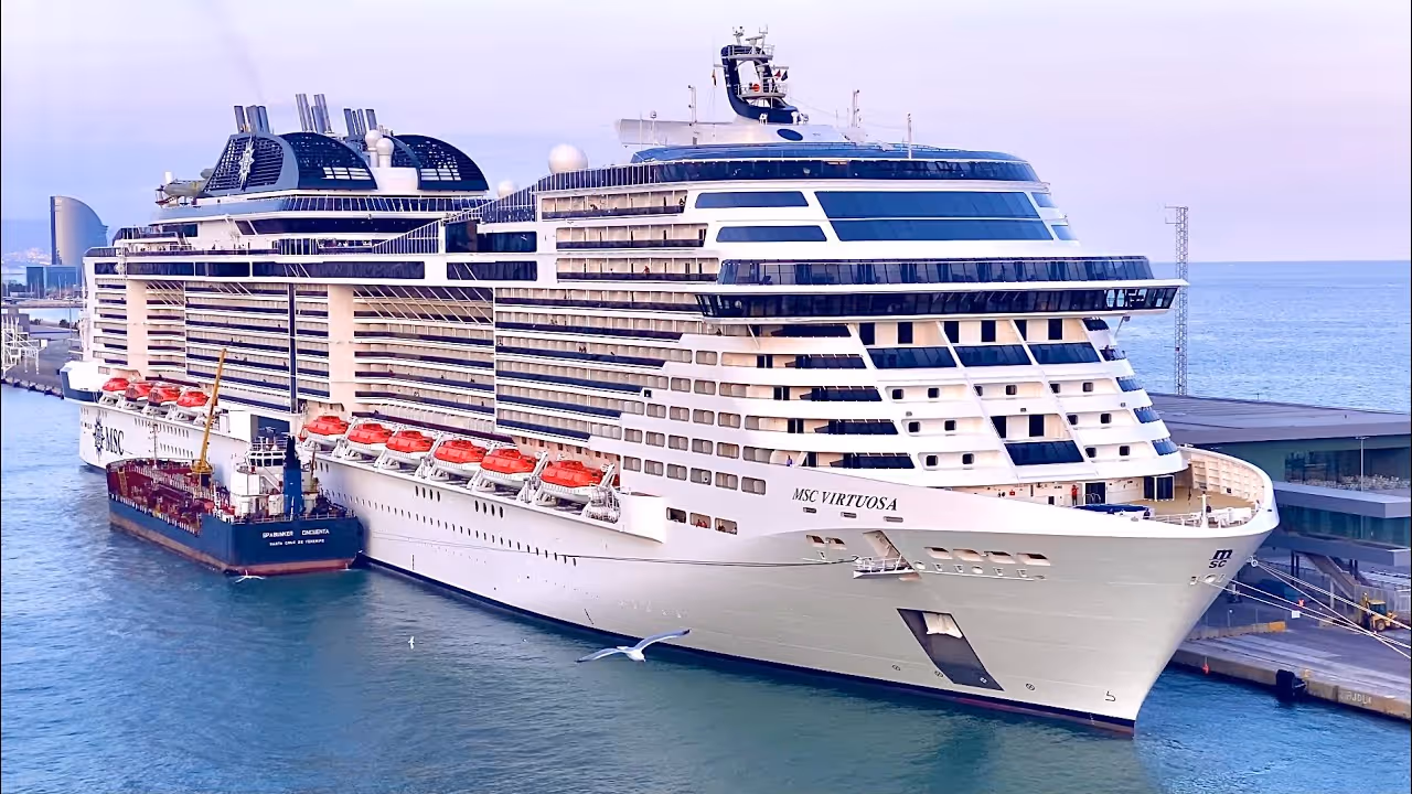 Large MSC Virtuosa cruise ship docked at a port with a smaller vessel alongside in calm water.