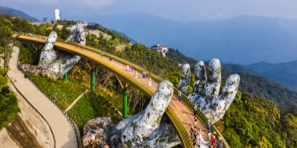 Elevated walkway supported by two giant sculpted hands amidst green hills with visitors walking along it.