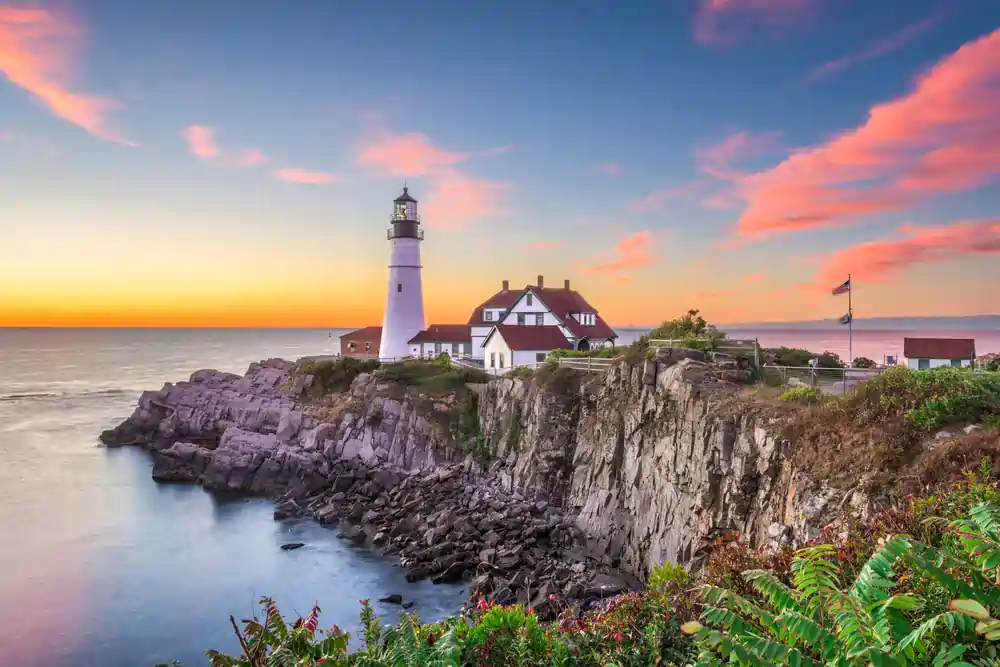 Lighthouse and buildings on rocky cliff overlooking calm ocean during colorful sunset.