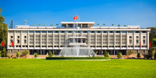 Modern government building with a central fountain and a flag on top, surrounded by a green lawn.