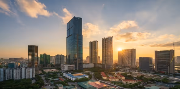 City skyline at sunset with modern high-rise buildings and sun shining through clouds.