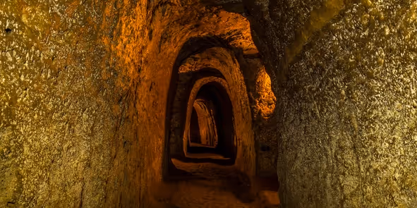 Dimly lit narrow tunnel with rough stone walls and arched ceilings extending into darkness.