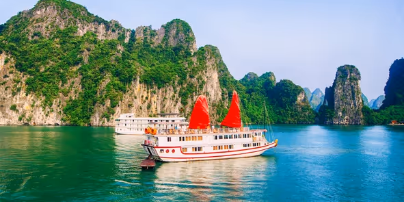 Traditional white cruise boat with red sails on calm blue water surrounded by green limestone cliffs.