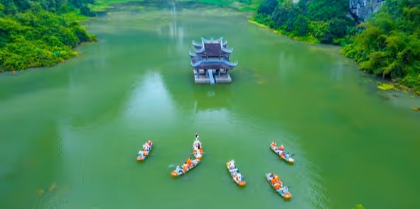 Aerial view of seven small boats with people rowing on a green river with a traditional pavilion in the center.