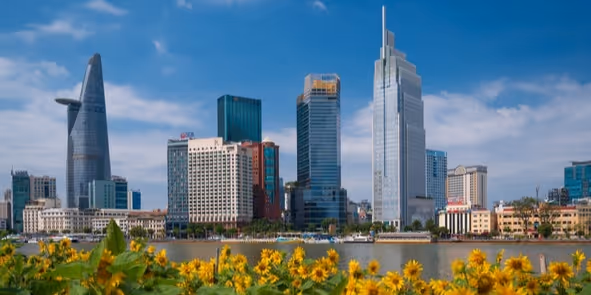 Skyline of modern skyscrapers along a river with yellow flowers in the foreground under a blue sky.