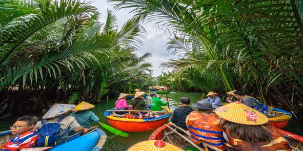 People wearing conical hats riding colorful basket boats along a narrow waterway lined with dense palm trees.