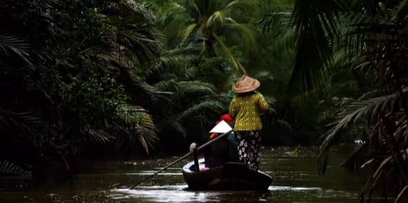 Two people wearing traditional hats paddling a small boat through a narrow river surrounded by dense tropical vegetation.