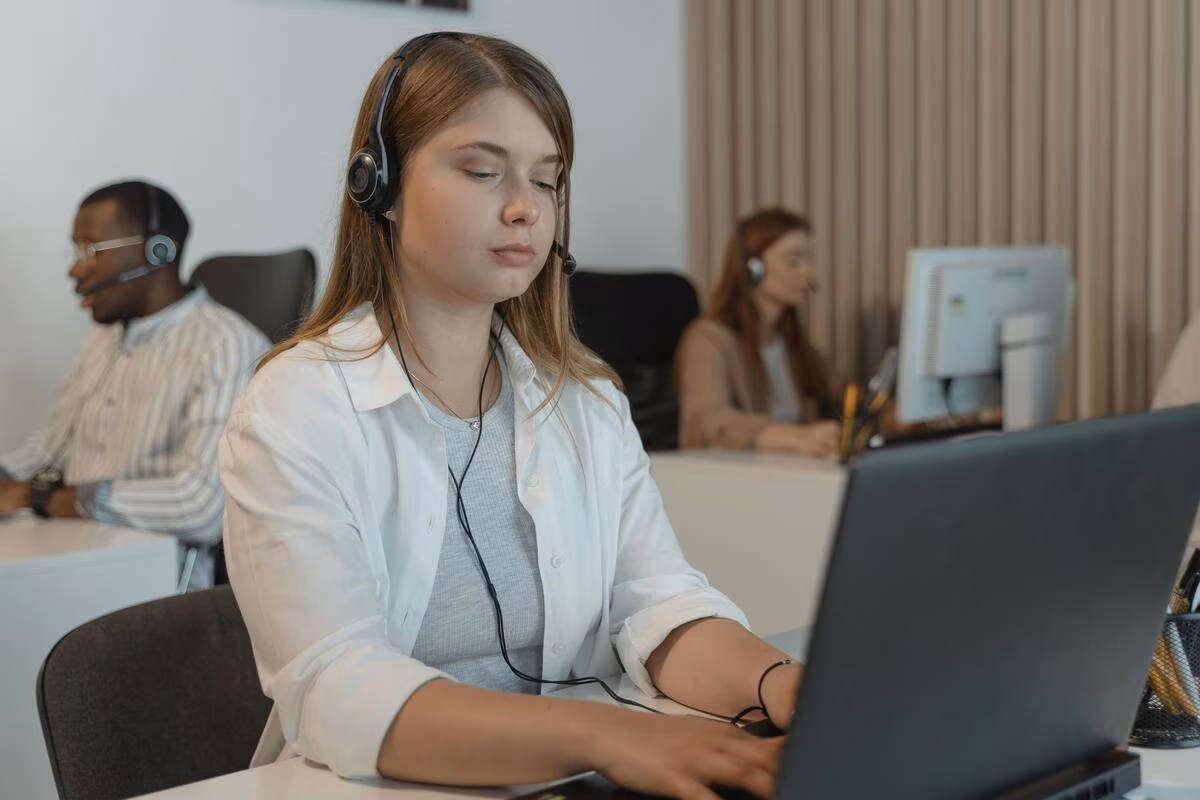 Woman wearing headset focused on typing on a laptop in a busy office with other call agents.