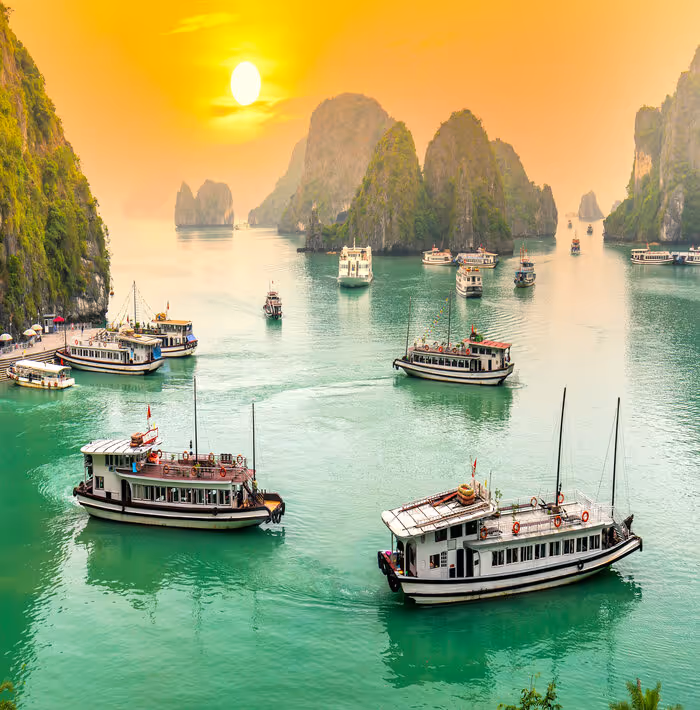 Several boats sailing in emerald green water among limestone islands at sunset in a scenic bay.